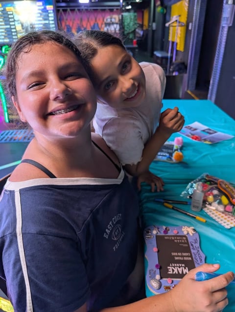 Two smiling children posing at a table with art supplies and gaming arcade machines in the background