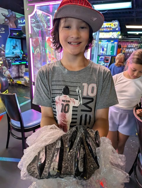 Smiling child wearing a gray soccer shirt and red cap holding a bucket of fried food at an arcade with colorful games in background