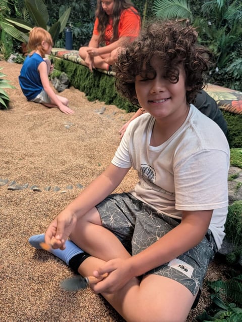 Child with curly hair smiling at camera while sitting on sandy ground during outdoor activity with others in background