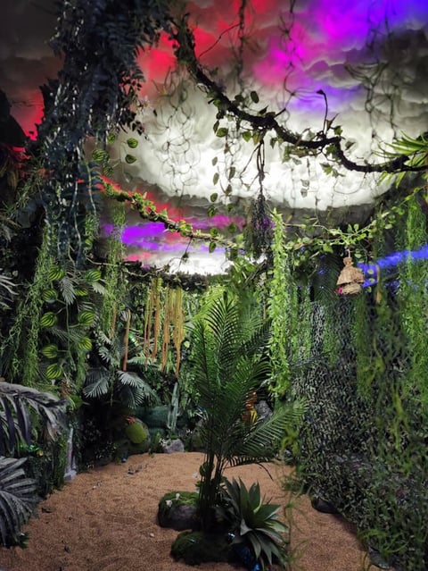 Indoor jungle installation with lush green plants and vines under dramatic colored lighting of pink, purple, and blue