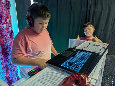 Two boys at a DJ booth with glowing keyboard in a studio with pink and teal neon lighting