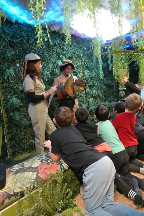 Zookeeper educating children about an animal in a lush, green indoor jungle exhibit with overhead lighting